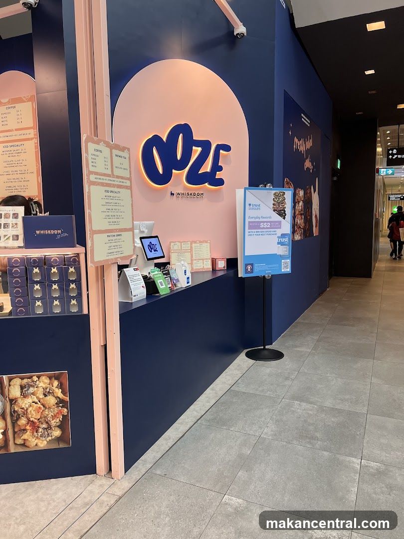 The shop front features a dark blue wall with a large pink arch displaying the neon sign "OOZE" in blue letters, with "BY WHISKDOM" written below it. To the left, a menu board lists coffee and iced specialty drinks. To the right, a stand displays a promotion for "Everyday Rewards" offering S$2 off. The counter area has a tablet and various payment devices.