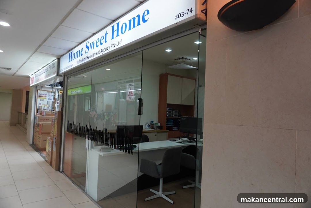 The exterior of a recruitment agency with a glass storefront. The sign above the entrance reads "Home Sweet Home Professional Recruitment Agency Pte Ltd" and the unit number "#03-74". Inside, a reception desk with computers and office chairs is visible.