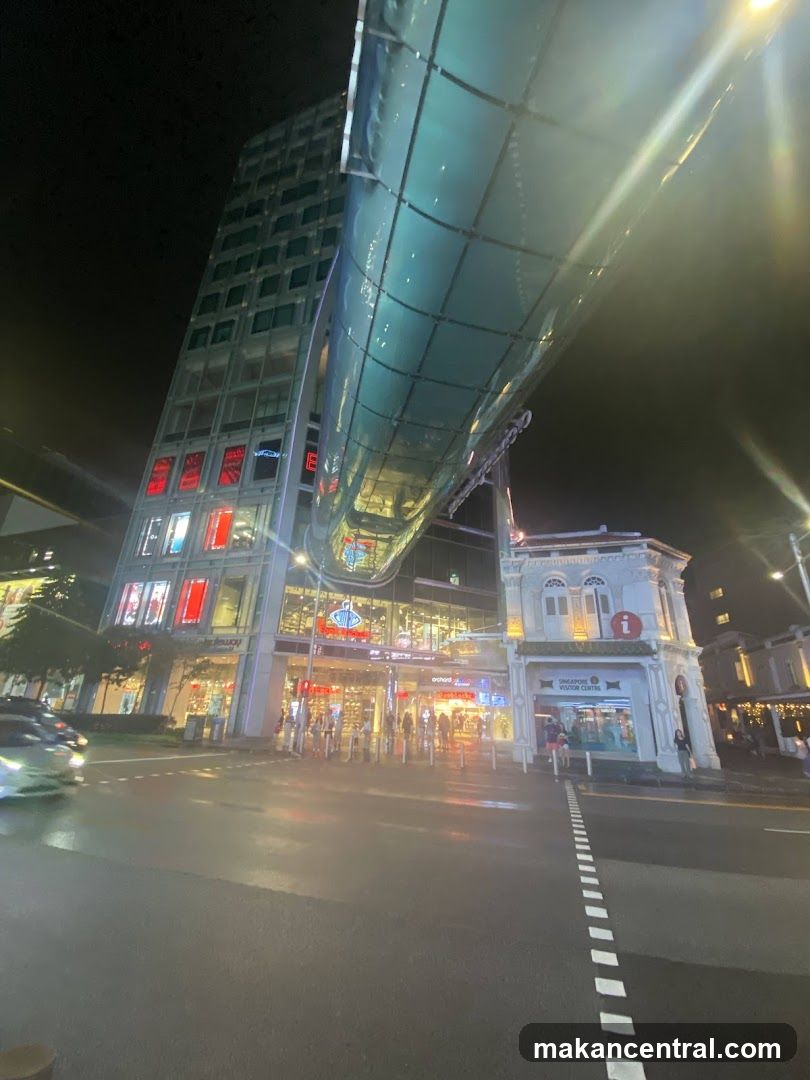 A nighttime view of a modern building with a glass walkway connecting to an older, ornate structure. The facade of the modern building features multiple levels of windows, some illuminated with red displays. The older building on the right has a sign that reads "SINGAPORE VISITOR CENTRE". The street in front is wet, reflecting the lights from the buildings.