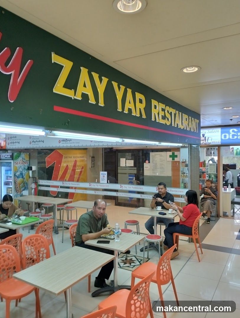The exterior of Zay Yar Restaurant, a dining establishment with a prominent green facade featuring the restaurant's name in large yellow letters. The interior is visible through large glass windows, showing tables and chairs set up for customers. Several patrons are seated and dining.