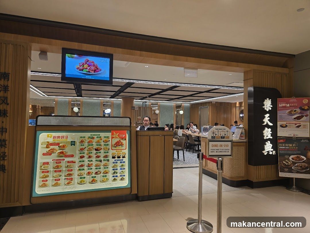 A view of a restaurant entrance with a large menu display on the left and a vertical sign with Chinese characters on the right. Inside, diners are seated at tables. A screen above the entrance displays food.