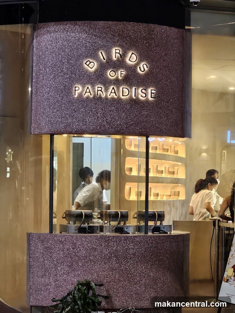 The exterior of a shop with the name "BIRDS OF PARADISE" illuminated in large letters above a glass display. Inside, staff members are visible preparing food, and shelves with cups or bowls are displayed.