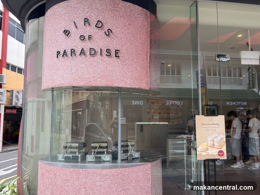 The exterior of a shop with a pink speckled facade and large glass windows. The shop's name, "BIRDS OF PARADISE," is displayed in metallic letters on the pink curved section. Inside, waffle makers are visible through the window. A sign on an easel outside advertises a "Party Cup Medley."