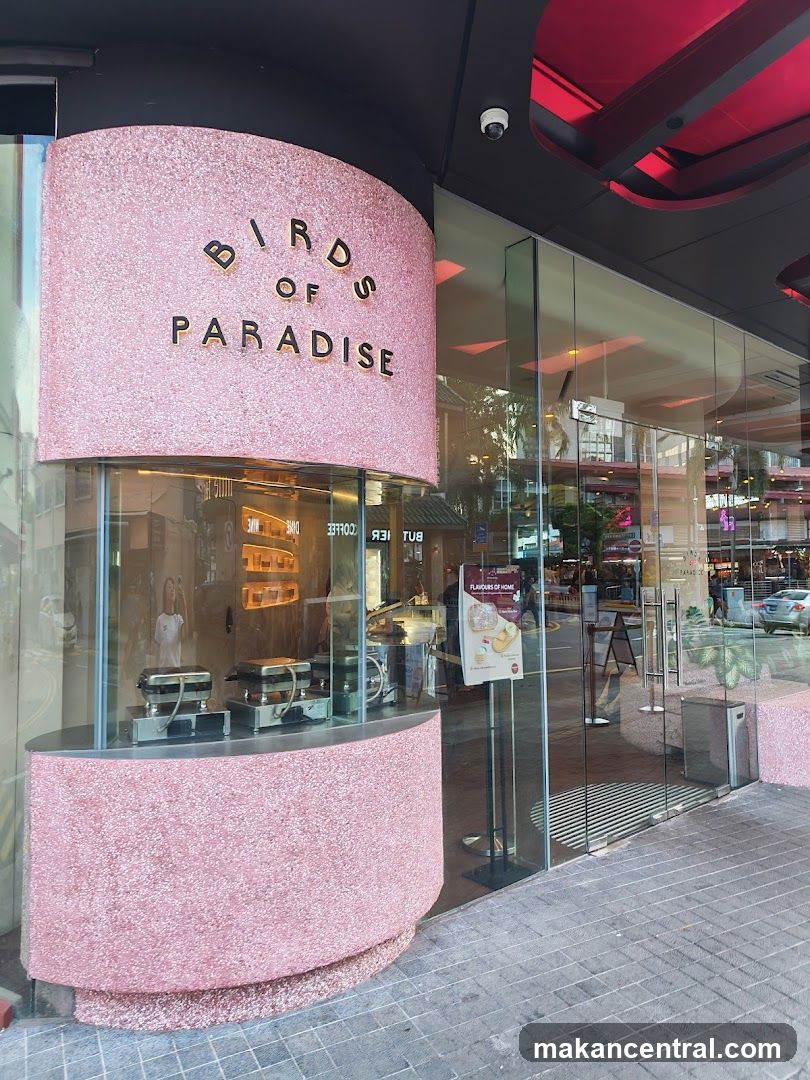 The exterior of a shop called Birds of Paradise. The shop has a pink, textured facade with the name displayed in gold lettering. Large glass windows reveal the interior, which appears to be a cafe or bakery with display cases and seating.