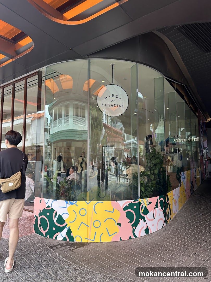 A modern shop front with large glass windows and a circular sign that reads "BIRDS OF PARADISE". The lower part of the windows is decorated with a colorful, abstract pattern. Inside, people are visible sitting at tables.