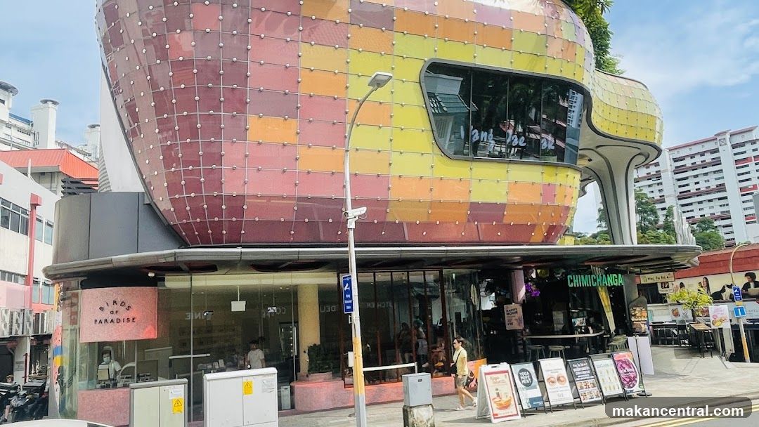 A storefront with a pink sign that says "BIRDS OF PARADISE" in black lettering. The shop has large glass windows and doors, and there are people inside. The building has a unique, colorful, curved facade above the storefront.