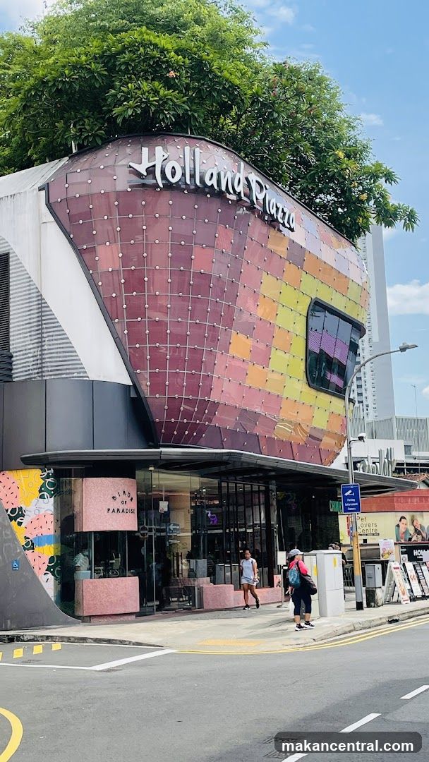 The exterior of a modern building with a curved facade covered in colorful square tiles. The building has a prominent sign that reads "Holland Plaza" at the top. Below the sign, there is a storefront with glass windows and doors. A pink structure with the words "BIRD OF PARADISE" is visible near the entrance. People are walking on the sidewalk in front of the building.