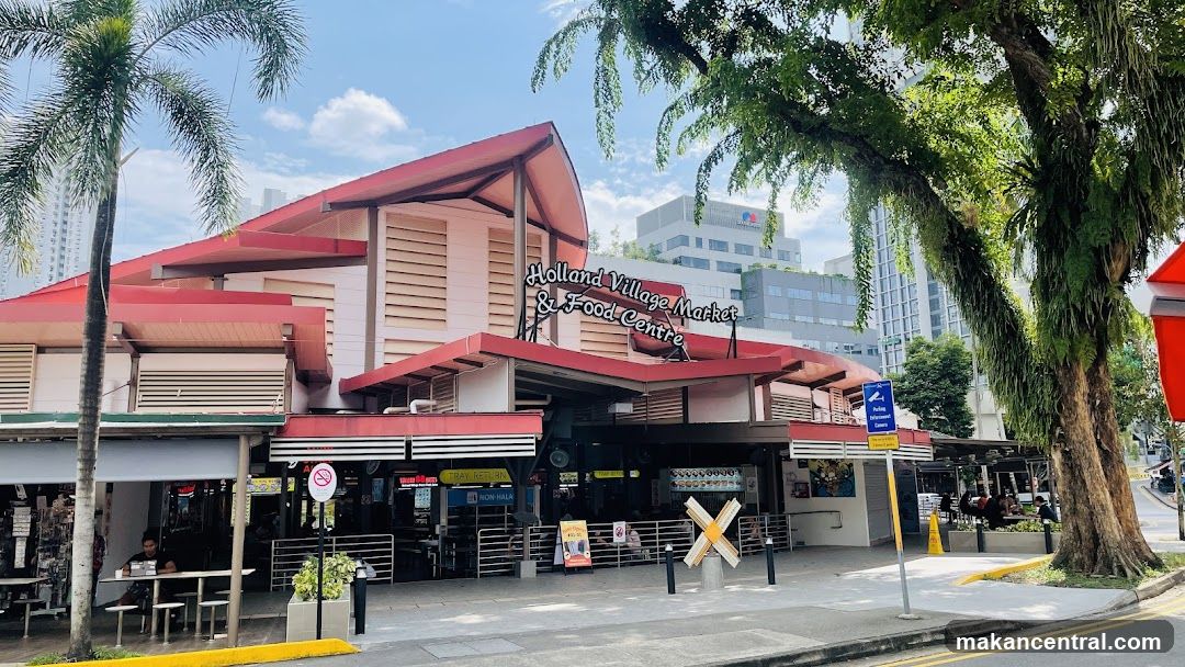 The exterior of the Holland Village Market & Food Centre building on a sunny day. The building has a red roof and white walls, with several stalls and seating areas visible. Palm trees and other greenery surround the building.