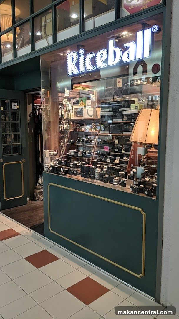 The exterior of a shop called "Riceball" is visible, featuring a large glass window displaying various cameras and camera equipment. The shop's name is illuminated in large, marquee-style letters above the window. To the left, a dark green door with gold trim is partially open, revealing racks of clothing or accessories inside. The interior of the shop appears to be well-lit, with shelves stocked with numerous camera lenses and bodies. A vintage-style lamp with a warm glow sits on a display shelf. The floor in front of the shop has a checkered pattern of cream and reddish-brown tiles.