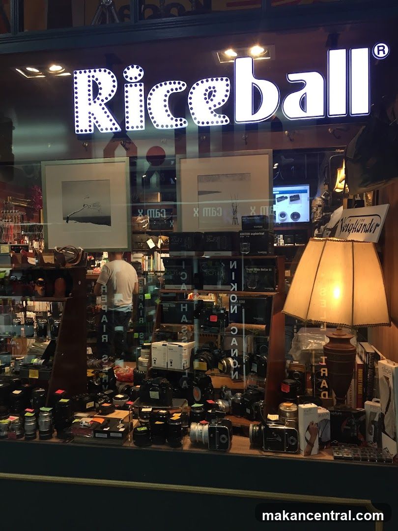 The storefront of a shop named Riceball, featuring a large illuminated sign. The window display showcases a variety of cameras, lenses, and photography equipment. Framed pictures hang on the wall behind the display. A lamp and books are also visible.