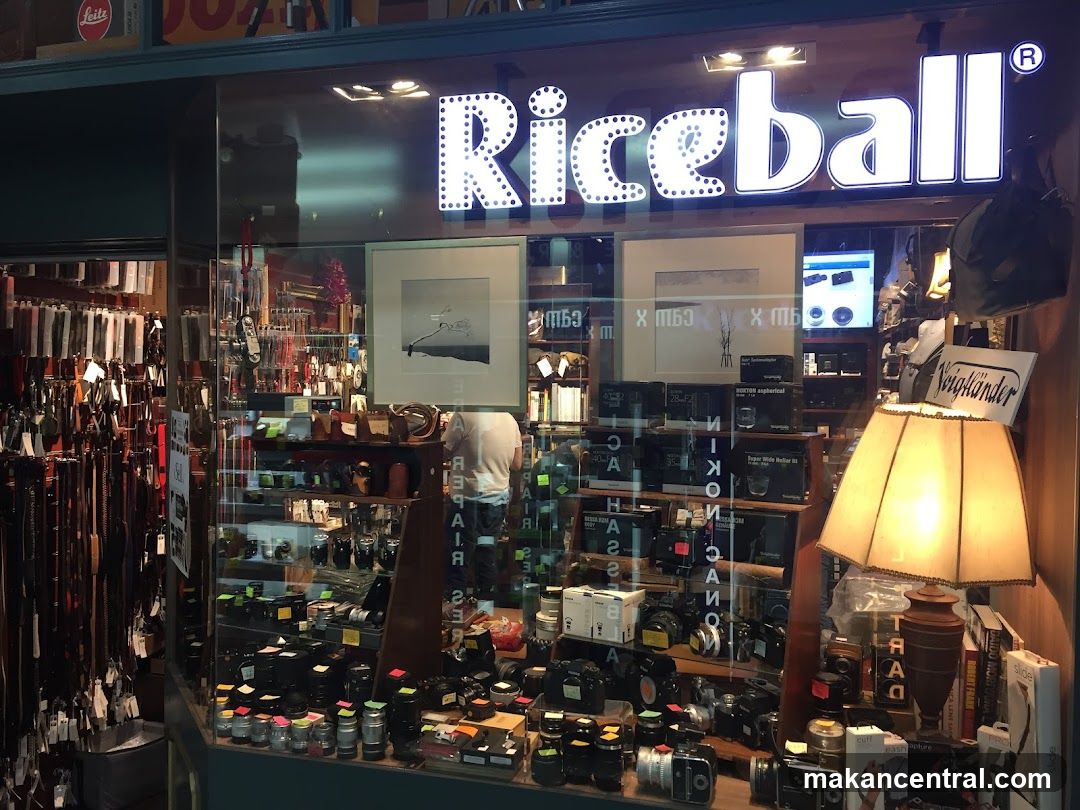 The storefront of a camera shop named Riceball. The shop window displays a variety of cameras, lenses, and camera accessories. There are also framed photographs and a vintage lamp visible in the display. The shop's name is illuminated in large letters above the window.