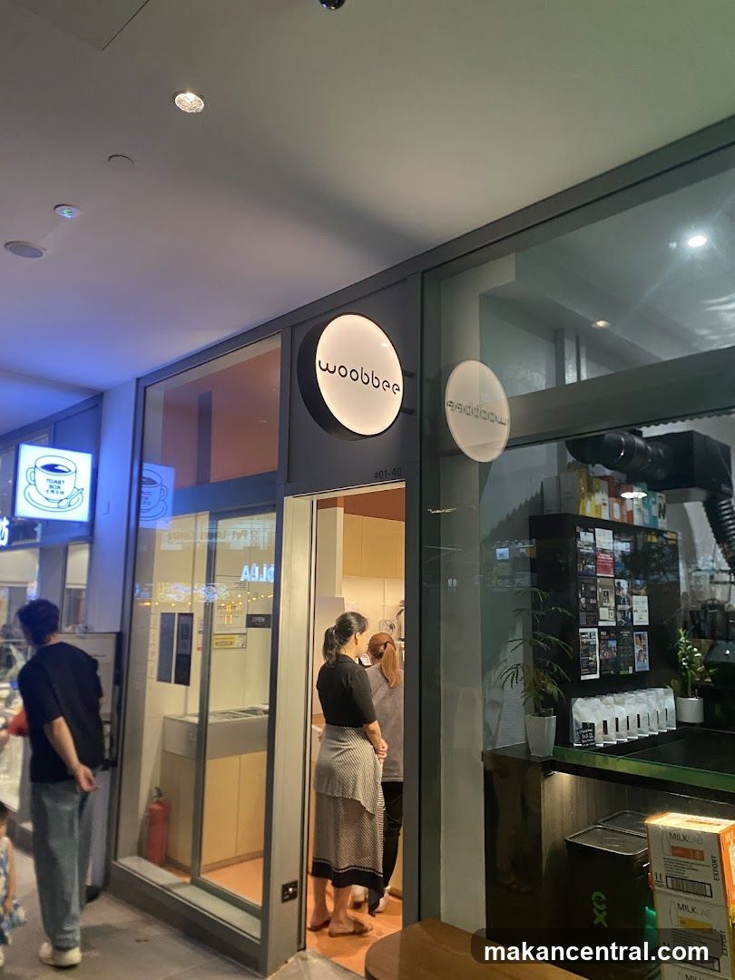 A view of the exterior of a shop with the name "woobbee" displayed on a circular illuminated sign. The shop has large glass windows and an open doorway. Inside, staff members are visible behind a counter.