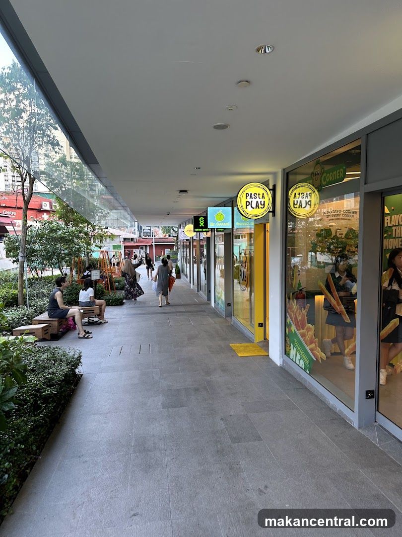 A row of shops with illuminated signs. The sign for "Pasta Play" is prominently displayed in a yellow circle. Another sign reads "Potato Corner". People are walking along the tiled walkway in front of the shops. There are also some people sitting on benches and lush greenery on the left side of the image.