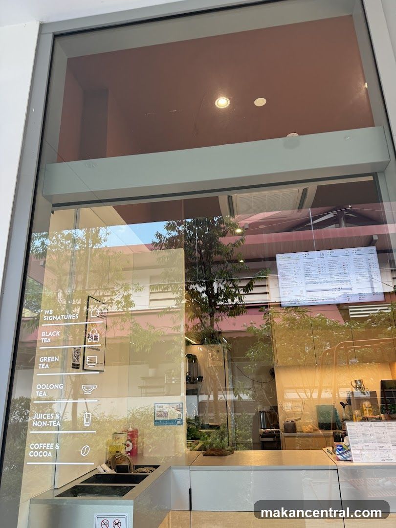 A view through a large glass window of a tea shop. On the left side of the window, a vertical list of beverage categories is displayed: WB SIGNATURES, BLACK TEA, GREEN TEA, OOLONG TEA, JUICES & NON-TEA, and COFFEE & COCOA. Each category has a corresponding icon. Inside the shop, a counter with a coffee machine and various items is visible. A menu board is displayed on the right side of the window.