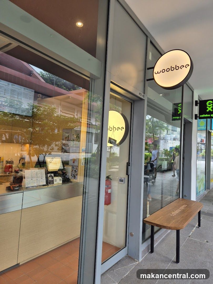 The exterior of a Woobee shop with a prominent circular sign displaying the logo. The shop has large glass windows and doors, revealing a counter and some interior elements. A wooden bench with the text "caffeine xpress" is placed outside the entrance.
