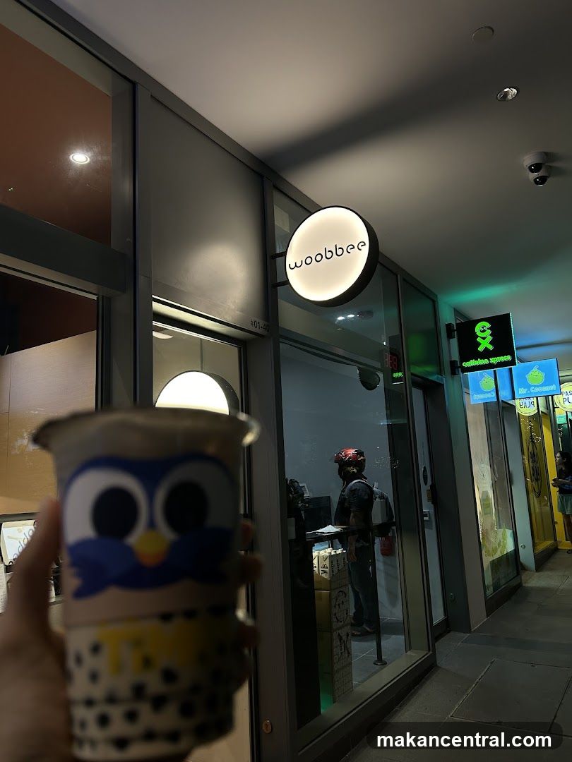 A person holds a bubble tea cup with a cartoon owl design in the foreground. In the background, the illuminated circular sign of 'woobbee' is visible, along with other shop signs and the interior of the store. A person wearing a helmet is seen inside the store.