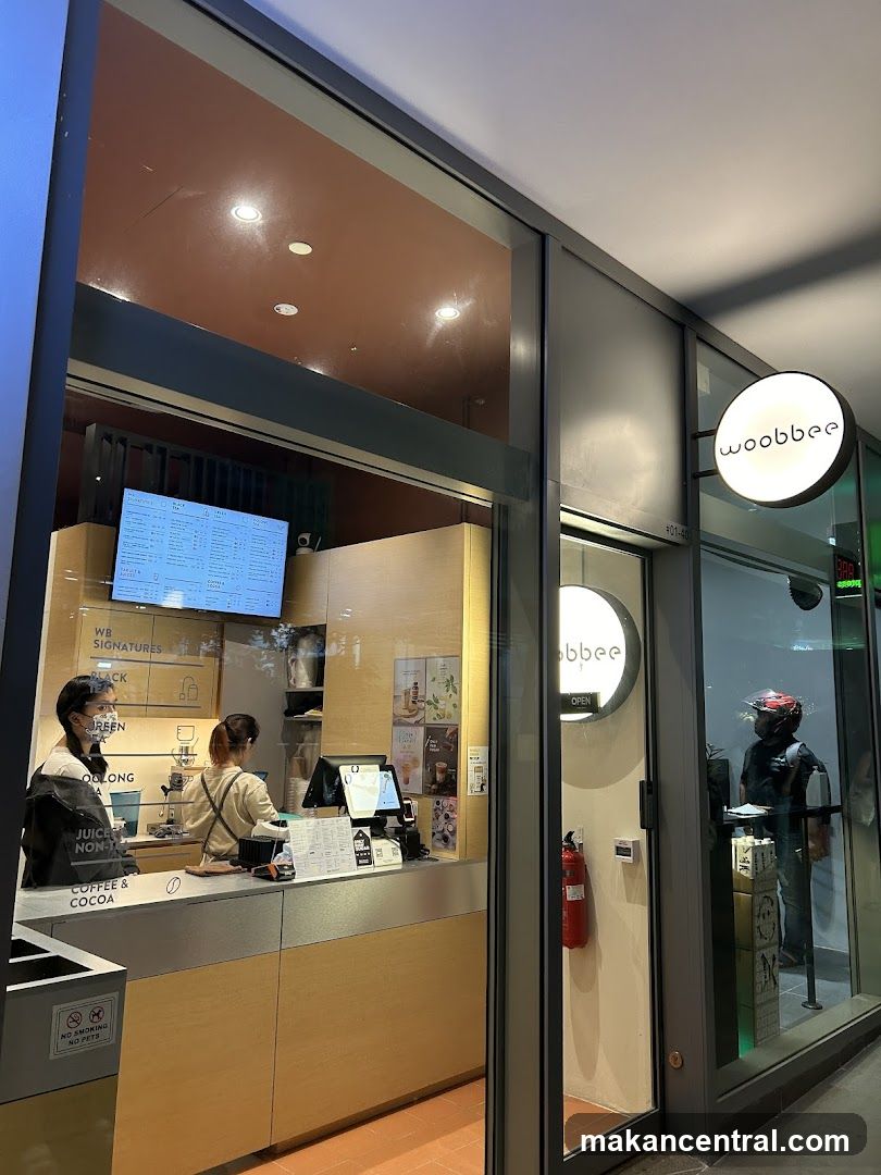 A view of the Woobbee store entrance and counter area. The store has a modern design with a large glass facade. A circular illuminated sign with the Woobbee logo is visible outside the entrance. Inside, a counter is staffed by two employees. A menu board is displayed above the counter.
