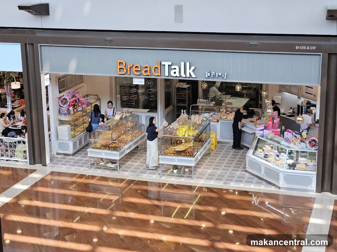 A BreadTalk bakery shop front with displays of bread, pastries, and cakes. Several customers are browsing and purchasing items. Staff members are visible behind the counter and in the kitchen area.