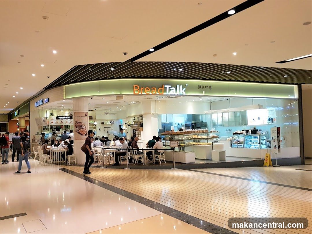 A BreadTalk bakery and cafe is shown from the outside, with customers seated at tables inside and a display of baked goods. The store has a modern design with a prominent "BreadTalk" sign. Adjacent to it is a "Toast Box" establishment.