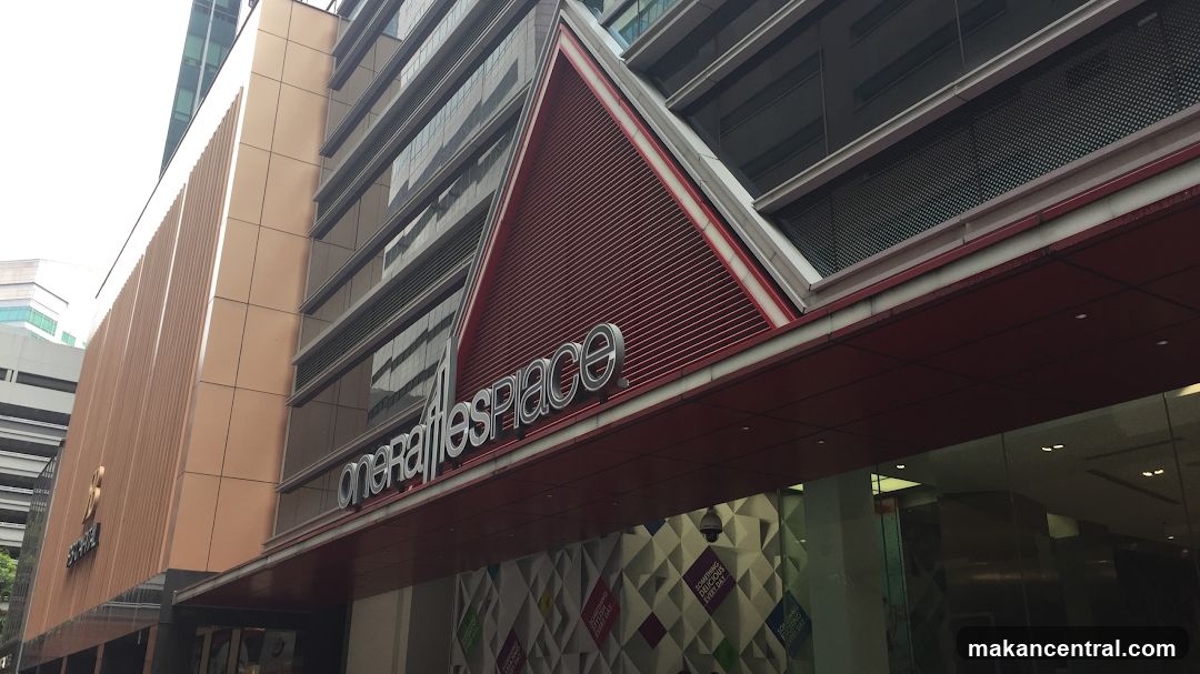 A close-up, low-angle shot of the exterior of the One Raffles Place building. The building features a modern design with metallic and glass elements. A prominent sign with the building's name, "one Raffles Place," is visible above the entrance. The lower part of the building has a patterned facade with text that reads "SOMETHING DELICIOUS EVERY DAY."