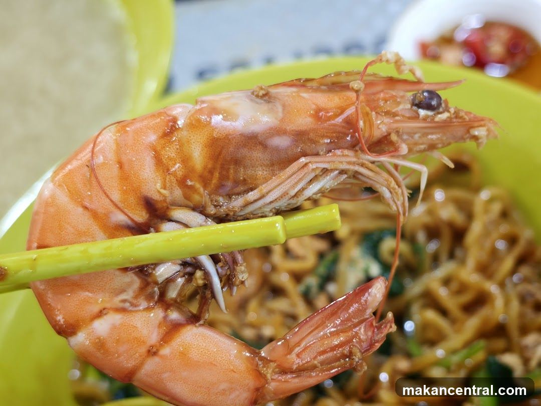 Close-up of a large prawn being held by chopsticks over a bowl of noodles.