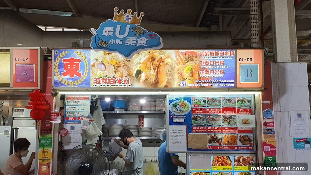 A food stall with a large illuminated sign displaying images of seafood white mee hoon and various dishes. The sign also lists menu items and prices. Below the main sign, there are smaller menu boards with pictures of food and prices. Staff members are visible working behind the counter. Red lanterns hang to the left of the stall.