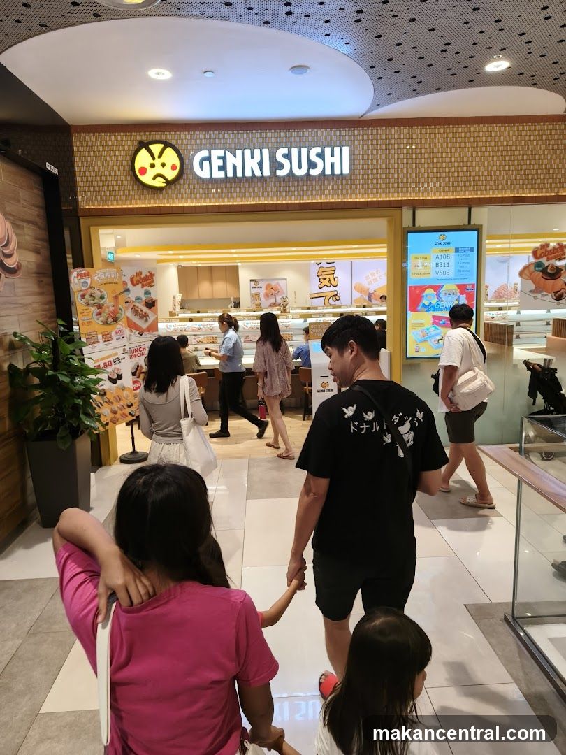 The exterior of a Genki Sushi restaurant with its prominent signage. Inside, staff are visible behind a counter, and customers are walking in. There are also promotional posters and a digital menu board displaying food items and estimated waiting times.