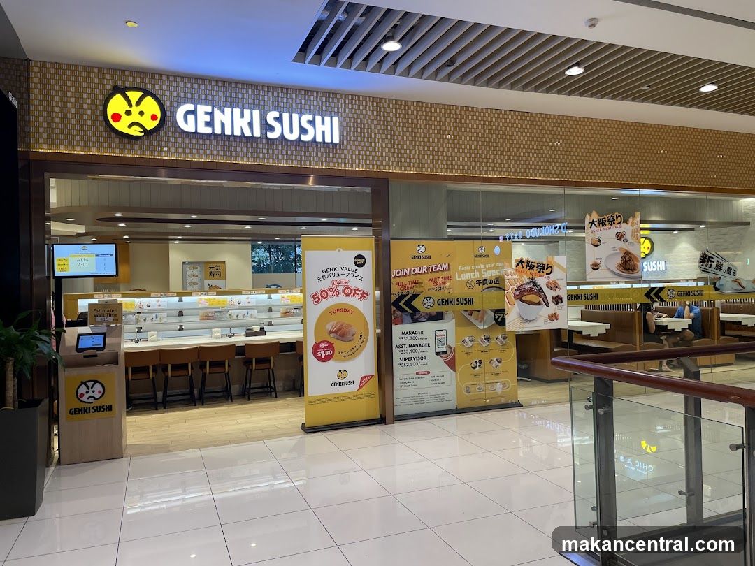 The exterior of a Genki Sushi restaurant with its prominent signage and a conveyor belt sushi bar visible inside. Promotional banners are displayed outside the entrance, advertising a 50% off Tuesday deal and job openings.