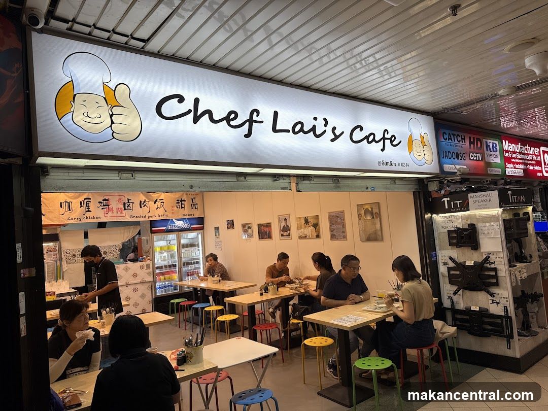 A brightly lit cafe with a large sign displaying the cafe's name and a cartoon chef giving a thumbs up. Below the sign, the cafe interior is visible with tables and chairs where customers are dining. A menu board with Chinese characters and English translations like "Curry chicken" and "Brasied pork belly" is displayed above the entrance.