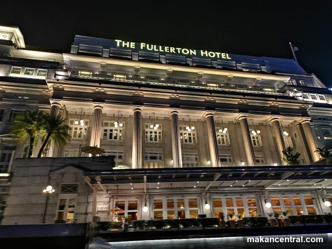 The exterior of The Fullerton Hotel at night, illuminated with warm lights. The hotel features a grand facade with classical columns and large windows. The hotel's name is prominently displayed in illuminated letters on the rooftop.