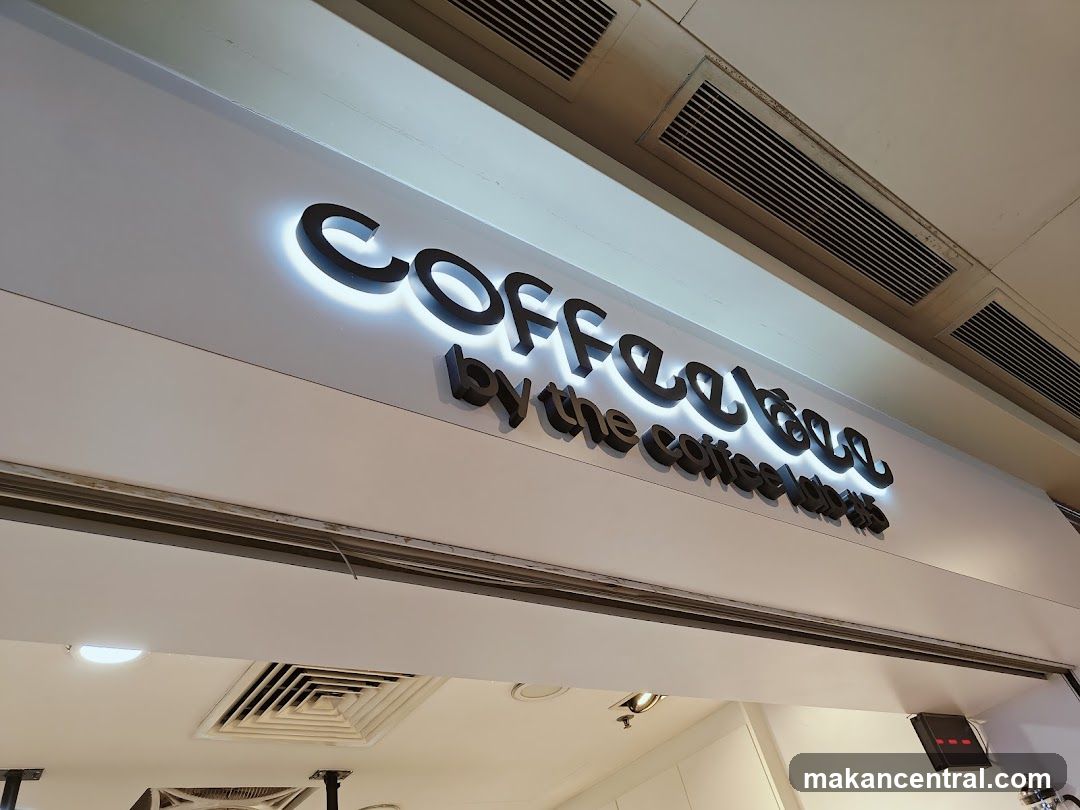 A close-up shot of the illuminated signage for "Coffee Bean by the coffee bean & tea leaf" mounted on a white facade. The letters are black and backlit with a soft white glow. Above the sign, ventilation grilles are visible on the ceiling. Below the sign, a dark horizontal strip runs across the facade, and a glimpse of the interior with lighting and a black box is visible.