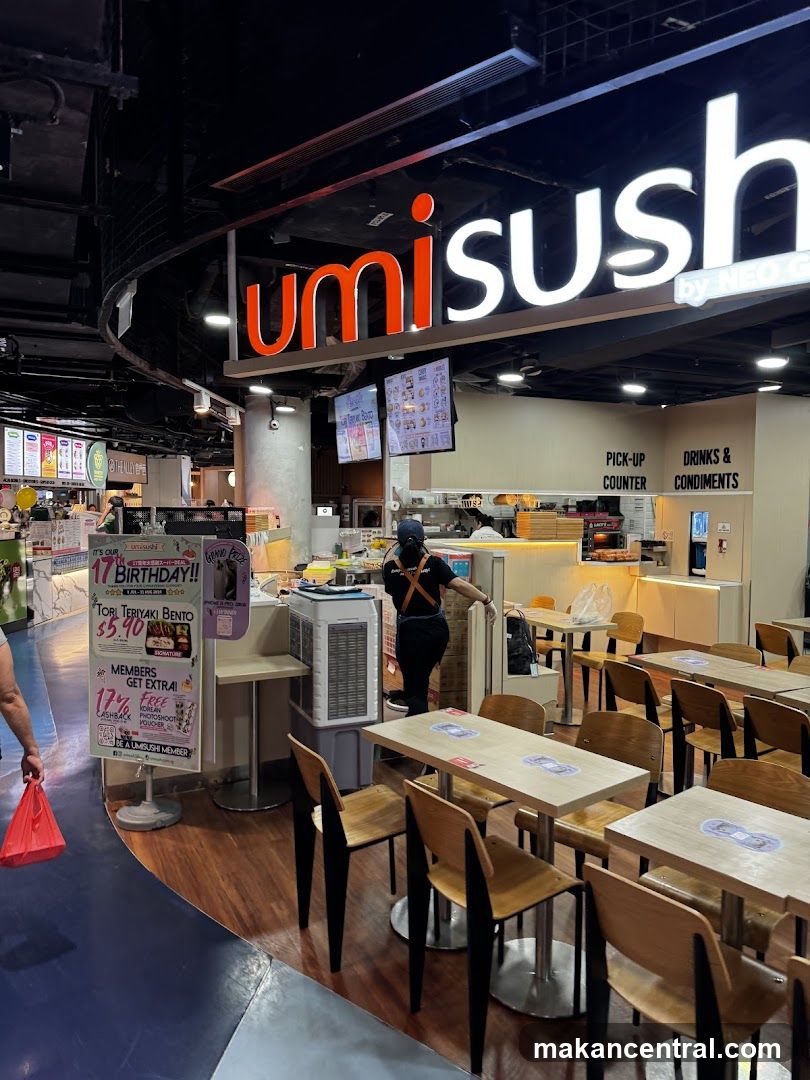 The exterior of a Umi Sushi restaurant with a prominent illuminated sign. The dining area with tables and chairs is visible, along with a pick-up counter and a drinks and condiments station. Promotional posters are displayed near the entrance.