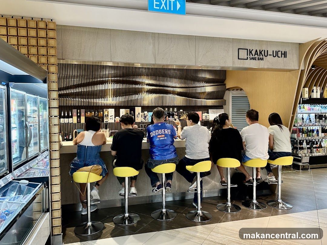 A group of people are seated at a bar in a sake bar. The bar has a modern design with wooden accents and a counter. There are bottles of sake displayed behind the counter. To the left, there are refrigerators with drinks. The sign above the bar reads "KAKU-UCHI SAKE BAR".