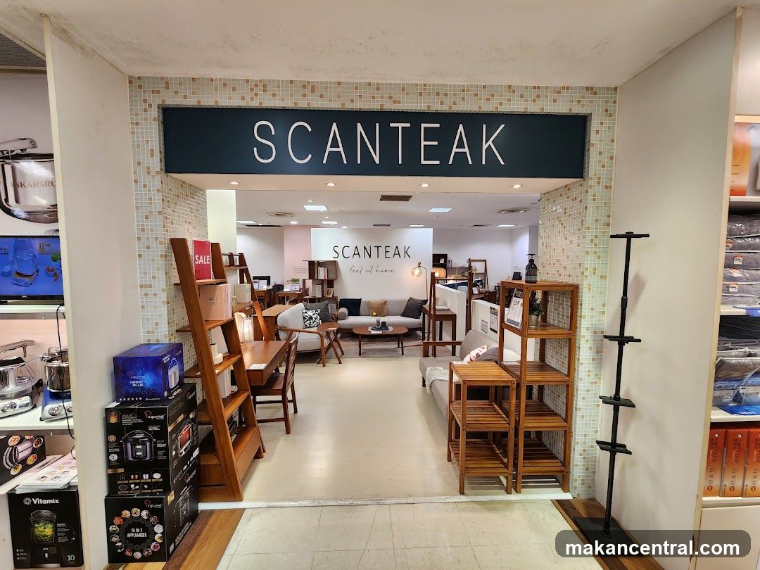 The entrance to a SCAN TEAK furniture store. The store name is displayed prominently on a dark blue sign above the entrance. Inside, various furniture items like sofas, chairs, and tables are displayed, along with shelving units and home decor. There are also some kitchen appliances and boxes stacked near the entrance.