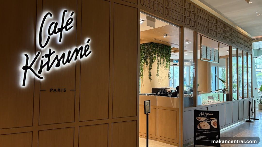 The exterior of a cafe with a prominent illuminated sign that reads "Café Kitsuné PARIS". The cafe has a modern wooden facade and large glass windows revealing the interior. Green vines cascade down a wall inside, and a counter with a coffee machine is visible. A menu board stands outside the entrance.