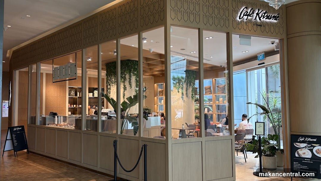 The exterior of a cafe with large glass windows and a prominent sign that reads "Cafe Kitsuné PARIS". Inside, shelves are stocked with merchandise, and plants hang from the ceiling. Customers are seated at tables. There are also A-frame signs outside with menu information.