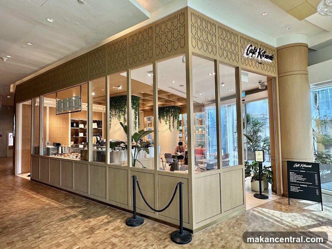 The exterior of a cafe with large glass windows and a wooden facade. The cafe's name, "Cafe Kitsuné," is displayed in illuminated lettering above the entrance. Inside, there are tables, chairs, plants, and displays of merchandise. A menu board is placed outside the cafe.