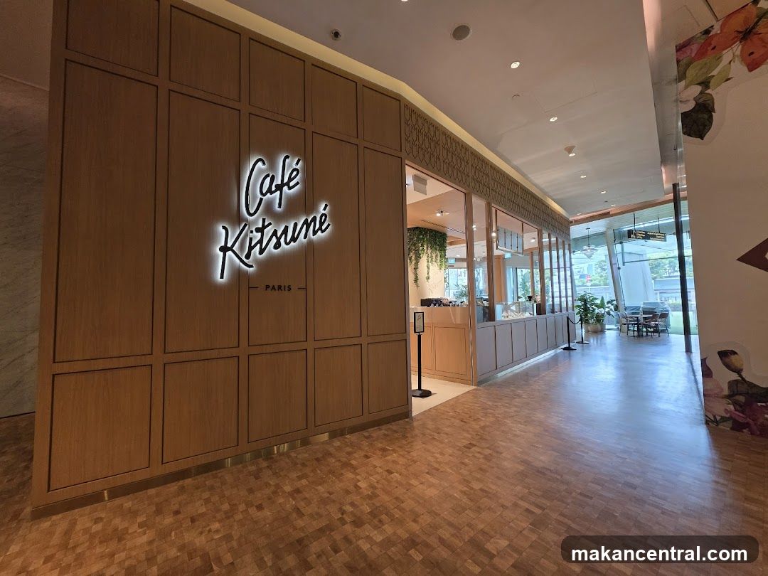 The exterior of Cafe Kitsune, a cafe with a prominent illuminated sign on a wooden paneled wall. The cafe counter and display cases are visible through glass windows.