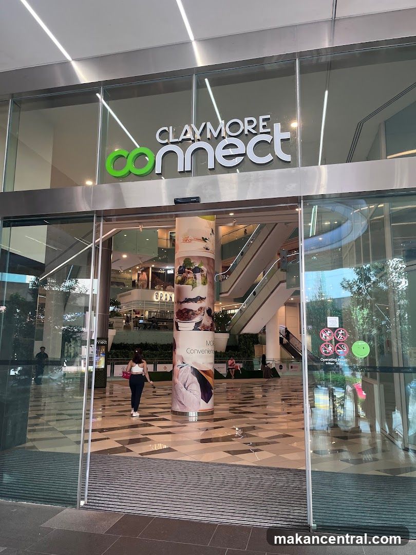 The exterior of the Claymore Connect building, featuring large glass doors and windows. The prominent signage "CLAYMORE connect" is visible above the entrance. Inside, escalators and various shops can be seen, along with a large cylindrical pillar displaying advertisements.
