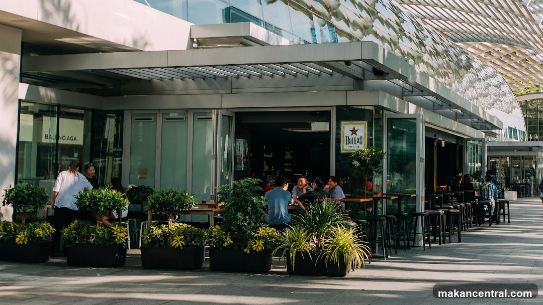 A street-level view of the Dallas Cafe & Bar storefront, featuring an open-air seating area with tables and chairs. People are seated at tables, some engaged in conversation. The cafe has large glass doors and windows, with planters filled with greenery lining the walkway in front. A sign with the cafe's name and logo is visible above the entrance. To the left, a Balenciaga store is also visible.