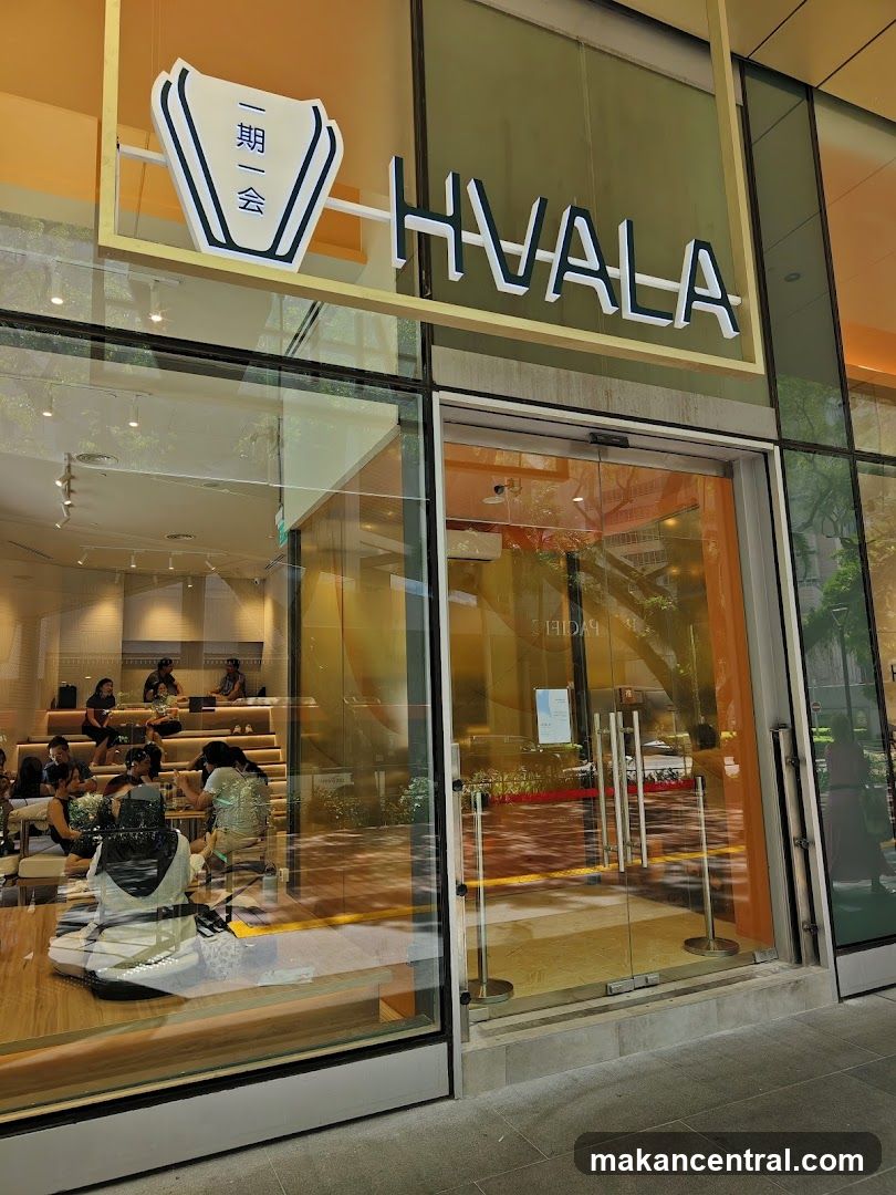 The exterior of a modern shop with large glass windows and doors. The shop's name, HVALA, is displayed in large, stylized letters above the entrance. Inside, tiered seating is visible with customers seated. The interior has a warm, inviting ambiance with wooden elements and soft lighting.