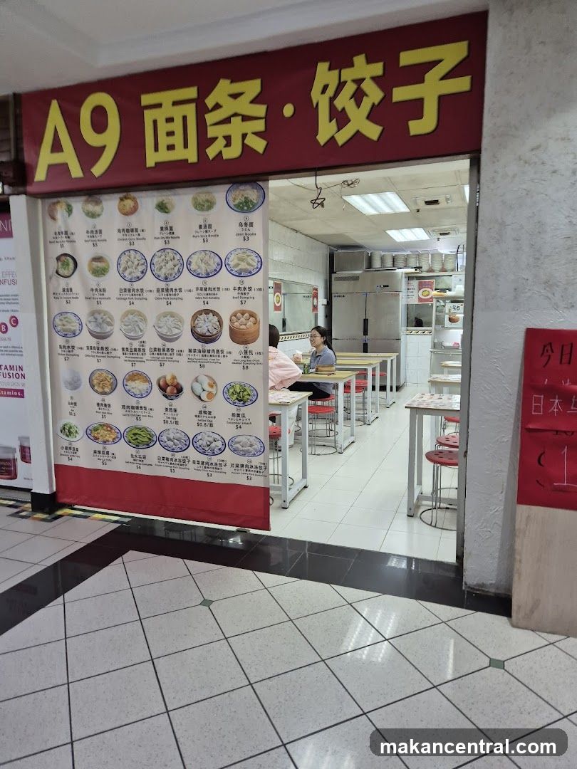 The storefront of a restaurant with a large red sign above the entrance displaying Chinese characters and "A9". Below the sign is a menu board with pictures and prices of various noodle and dumpling dishes. The interior of the restaurant is visible, showing tables, chairs, and kitchen equipment.