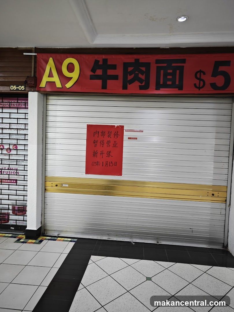 A closed shop with a red banner above the metal shutter door. The banner reads "A9 Beef Noodles $5" in yellow and black text. A red sign is posted on the shutter door, stating "Internal renovation, temporarily closed, new opening (25th year) January 15th". To the left, another shop's shutter is partially visible with some text and product images. The floor is tiled in a diamond pattern.