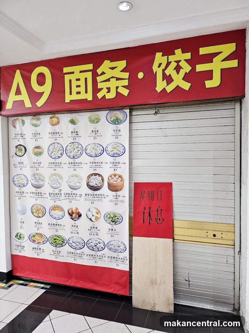 A shop front with a large red banner displaying "A9 面条・饺子" in yellow Chinese characters. Below the banner is a menu board showcasing various noodle and dumpling dishes with images and prices. To the right, a red and beige sign reads "星期日 休息" (Sunday Rest). The shop's metal shutter is partially down.