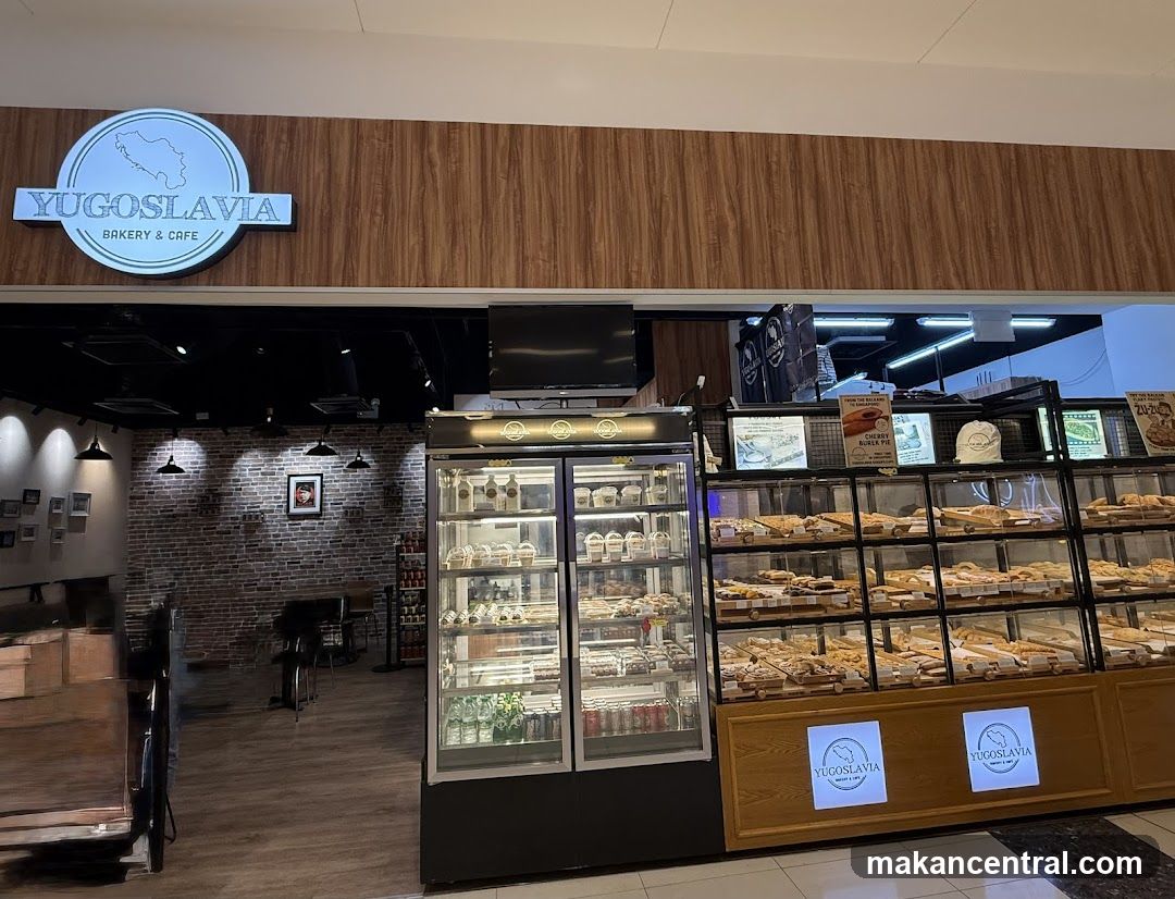 The exterior of a bakery and cafe with a prominent sign that reads "YUGOSLAVIA BAKERY & CAFE" inside a circular logo featuring a map outline. The storefront features a display of baked goods in glass cases and a refrigerated display. The interior visible through the entrance shows brick walls and seating areas.