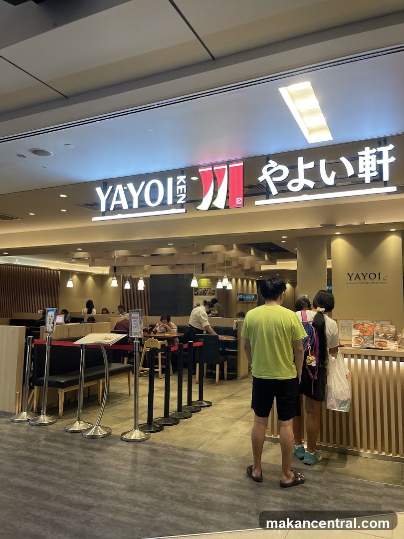 The exterior of a Japanese restaurant called Yayoi Ken. The restaurant has a large illuminated sign with the name in English and Japanese characters. Inside, diners are seated at tables, and staff members are visible. There are stanchions and signs indicating a waiting area.