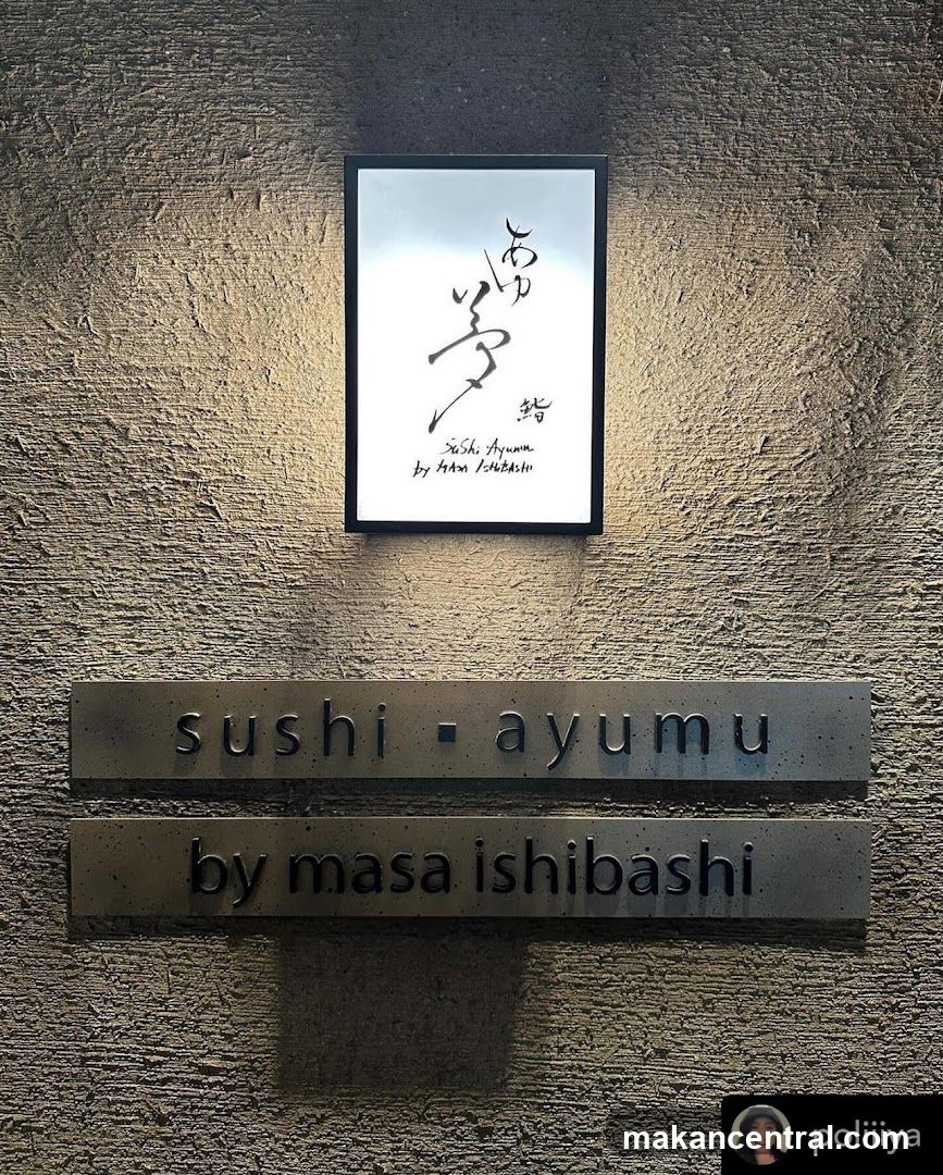 A sign for "sushi - ayumu by masa ishibashi" is mounted on a textured wall. Above the metal signs, a framed artwork with Japanese calligraphy and English text is illuminated.