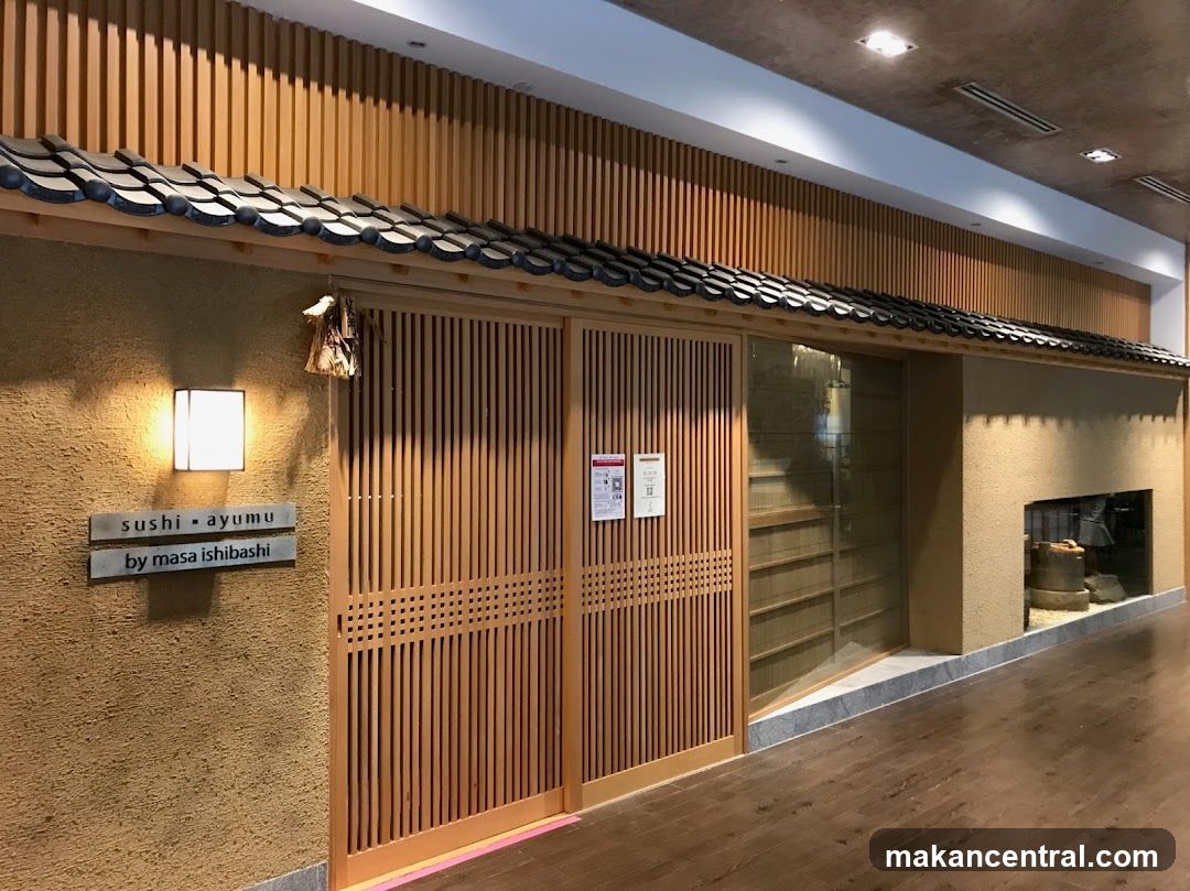 The exterior of a Japanese restaurant with wooden sliding doors and a textured beige wall. A sign reads "sushi ayumu by masa ishibashi". Traditional Japanese roof tiles are visible above the entrance.
