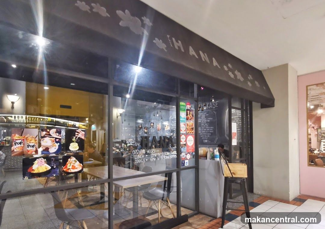 The storefront of a restaurant named HANA. The entrance features large glass windows and a dark awning with the restaurant's name and floral designs. Inside, tables and chairs are visible, along with menu boards and displays of food items.