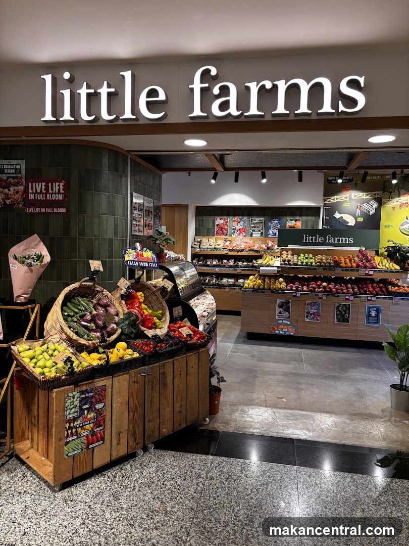 The exterior of a grocery store called Little Farms. The store has a large sign with the name "little farms" in white illuminated letters. In front of the store, there are wooden crates filled with fresh produce like eggplants, peppers, tomatoes, and pears. Inside the store, shelves are stocked with various fruits and vegetables.